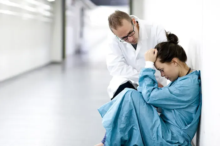 Kvinne i grøn sjukehusuniform sit på golvet i ein korridor og tek seg til hovudet. Ein mann i kvit frakk lener seg hjelpdamt over henne