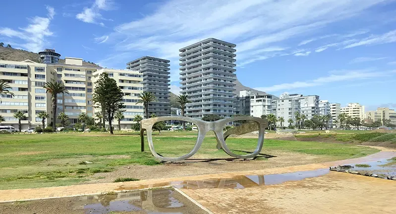 Ein skulptur som føresteller eit par meterhøge briller ved eit strandområde. Bustadblokker i bakgrunnen