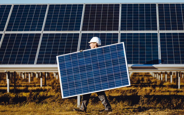 mann med hjelm bærer solcellepanel. i bakgrunnen solcellepark